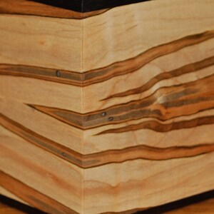 May include: A wooden box with a natural wood grain pattern. The box is light-colored with dark brown streaks and accents. The corners are joined with small metal rivets, and the base is dark.