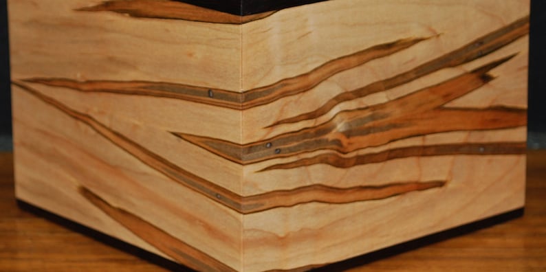 May include: A wooden box with a natural wood grain pattern. The box is light-colored with dark brown streaks and accents. The corners are joined with small metal rivets, and the base is dark.