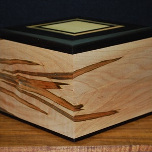 May include: A handcrafted wooden box with a square shape. The box features a light-colored wood with dark brown streaks and a black top. The top has a lighter square inlay. The box is sitting on a wooden surface.