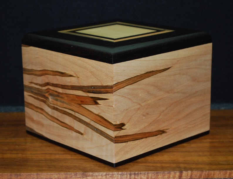 May include: A handcrafted wooden box with a square shape. The box features a light-colored wood with dark brown streaks and a black top. The top has a lighter square inlay. The box is sitting on a wooden surface.
