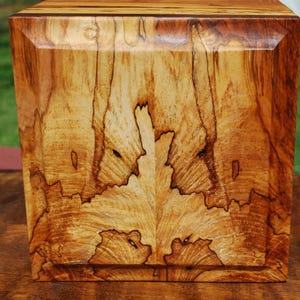 May include: A wooden box with a square shape and a natural wood grain pattern. The box is made of light brown wood with darker brown streaks and patterns. The edges of the box are beveled, and the wood grain is visible on all sides.