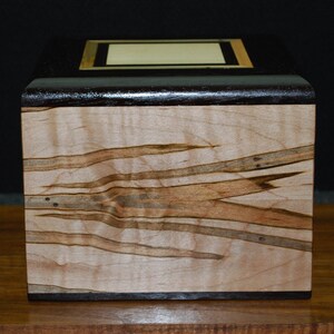 May include: A handcrafted wooden box with a dark brown top and a light-colored, patterned body. The top features a rectangular inlay with a gold border. The box is made of wood and has a smooth finish.