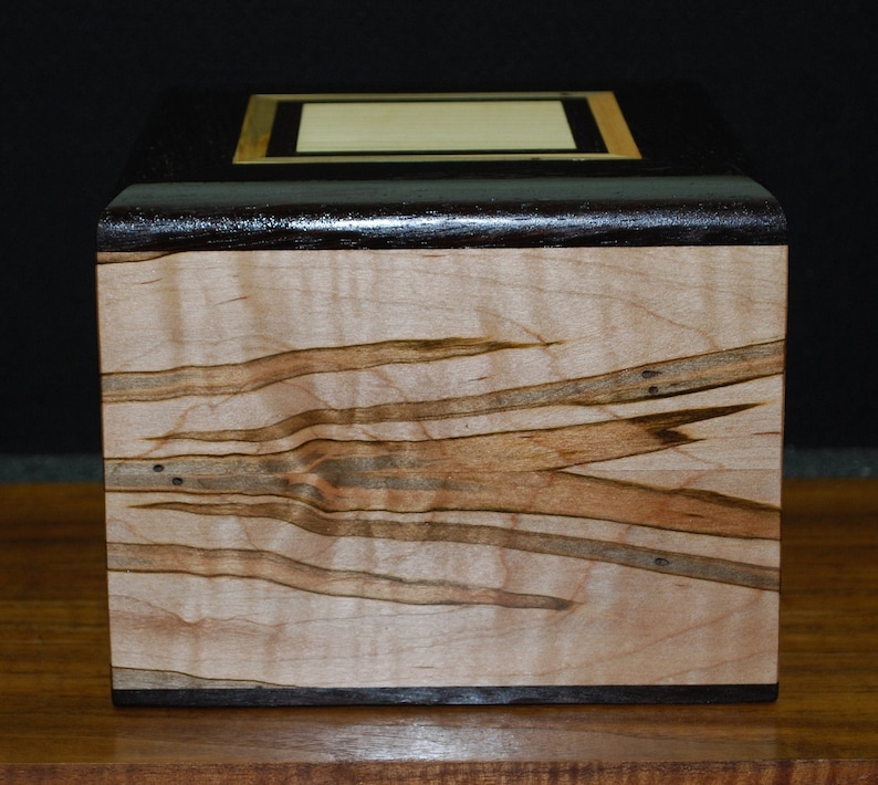 May include: A handcrafted wooden box with a dark brown top and a light-colored, patterned body. The top features a rectangular inlay with a gold border. The box is made of wood and has a smooth finish.