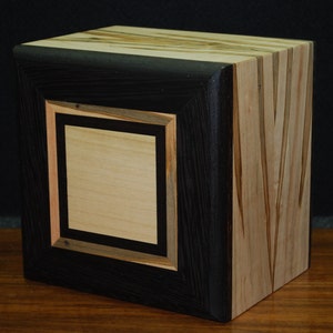 May include: A handcrafted wooden cube with a black frame and a light-colored center panel. The cube's sides feature a mix of light and dark wood grains, showcasing the natural patterns and textures of the material. The design is geometric and modern.