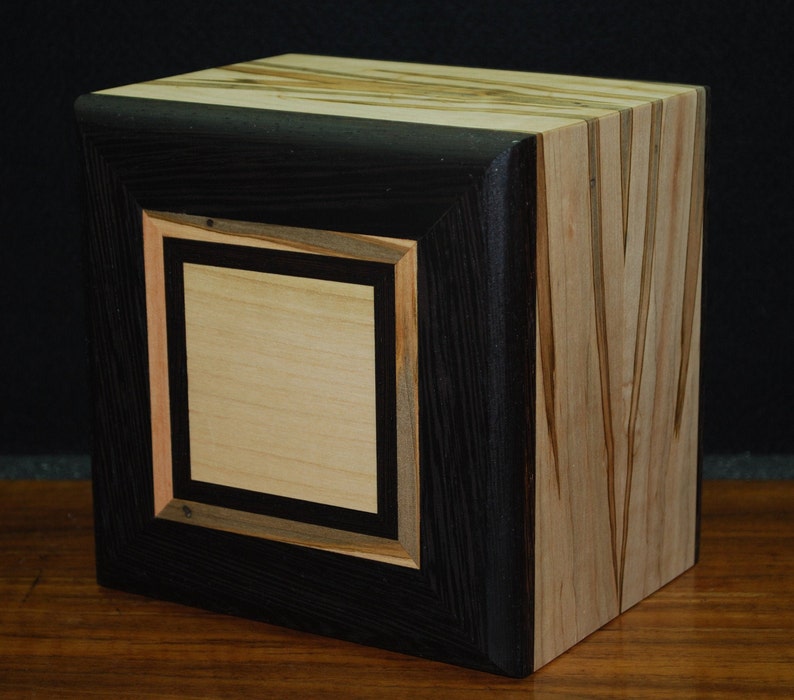 May include: A handcrafted wooden cube with a black frame and a light-colored center panel. The cube's sides feature a mix of light and dark wood grains, showcasing the natural patterns and textures of the material. The design is geometric and modern.