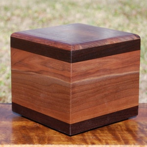Op de afbeelding: Een vierkante houten doos met een donkerbruin houten bovenkant en zijkanten, en een lichter bruin houten onderkant en zijkanten. De doos is gemaakt van walnoot hout en heeft een natuurlijke afwerking.