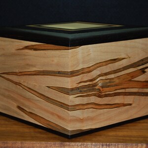May include: A wooden box with a light-colored wood grain pattern and dark brown accents. The box has a square shape with a flat top and a dark border. The wood grain has a unique, natural design.