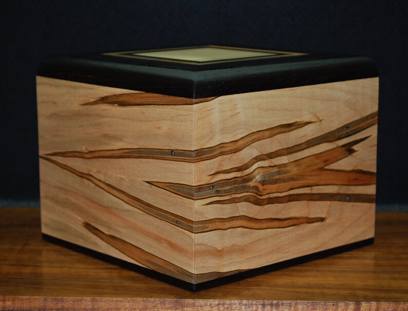 May include: A wooden box with a light-colored wood grain pattern and dark brown accents. The box has a square shape with a flat top and a dark border. The wood grain has a unique, natural design.