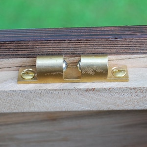 May include: Close-up of a brass door catch with two ball bearings. The catch is mounted on a wooden surface.