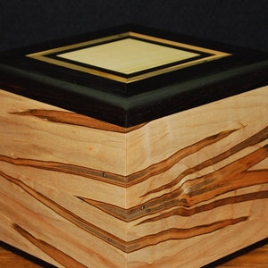 May include: A handcrafted wooden box with a geometric design. The box features a light-colored wood body with dark brown and tan streaks. The top is black with a light wood and gold-colored square inlay. The box is likely used for storage.