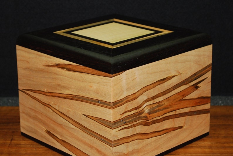 May include: A handcrafted wooden box with a geometric design. The box features a light-colored wood body with dark brown and tan streaks. The top is black with a light wood and gold-colored square inlay. The box is likely used for storage.