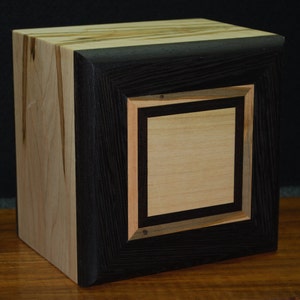 May include: A handcrafted wooden box with a square design. The box features a light-colored wood on the sides and top, contrasted by a dark, almost black, frame around a central square. The central square is a lighter wood, creating a layered effect.