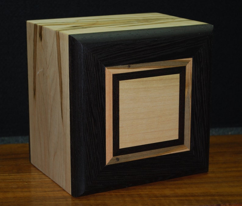 May include: A handcrafted wooden box with a square design. The box features a light-colored wood on the sides and top, contrasted by a dark, almost black, frame around a central square. The central square is a lighter wood, creating a layered effect.