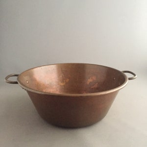 May include: A large, round copper bowl with two handles. The bowl has a worn, aged patina and is slightly dented.