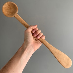 Könnte beinhalten: Ein langer Holzlöffel mit einer abgerundeten Schüssel und einem langen Stiel. Der Löffel wird in der Hand einer Person gehalten.