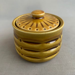 May include: A small, round ceramic jar with a lid. The jar and lid are a warm, mustard yellow colour with brown accents. The lid has a knob on top and a sunburst design. The jar has a textured, ribbed pattern with brown dots.
