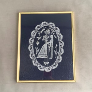 May include: A framed piece of white lace artwork depicting a couple standing together. The lace is intricate and detailed, and the couple is dressed in traditional clothing. The artwork is set against a dark blue background.