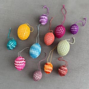 Könnte beinhalten: Eine Sammlung farbenfroher, gehäkelter Ostereier-Ornamente in verschiedenen Blau-, Gelb-, Rosa-, Orange- und Grüntönen. Jedes Ei hat eine Schlaufe zum Aufhängen. Die Eier sind auf einer grauen Oberfläche angeordnet.