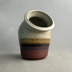 Può includere: Un vaso in ceramica fatto a mano con un'apertura angolata unica. Il vaso presenta una sfumatura di colore, che passa da un bordo bianco crema a fasce di beige, marrone e rosso intenso. La ceramica ha un aspetto rustico e strutturato.
