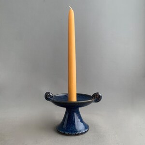 May include: A dark blue ceramic candle holder with a flared base and two curved handles. An orange taper candle is inserted in the center. The candle holder has a glossy finish and a decorative rim.