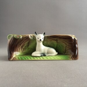 May include: A ceramic planter shaped like a tree trunk with a white fawn lying on a green leaf bed. The planter is decorated with brown, green, and white glaze.