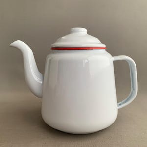 Puede incluir: Una tetera de esmalte blanco con borde rojo y tapa a juego. La tetera tiene un pico curvo y un asa. Este artículo de cocina clásico es perfecto para preparar té u otras bebidas calientes.