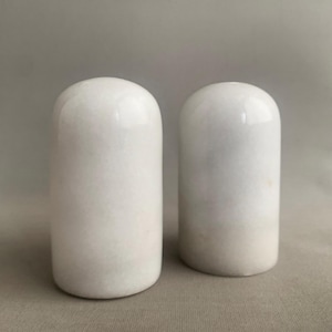 Puede incluir: Dos saleros y pimenteros blancos, cilíndricos, con tapas redondeadas. Los saleros y pimenteros tienen un acabado liso y brillante y un aspecto ligeramente texturizado. Se muestran sobre un fondo neutro.