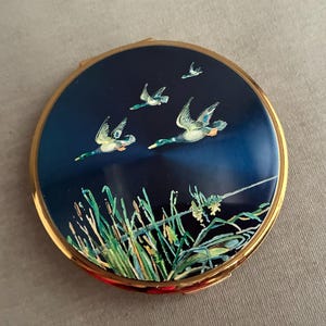 May include: A round, vintage compact with a gold-tone rim. The navy blue lid features a painted scene of flying ducks above reeds. The ducks are in flight, with green, white, and orange details. The compact is a classic accessory.