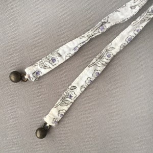 May include: Two white fabric cords with a floral pattern and purple accents. Each cord has a silver metal bead at the end.