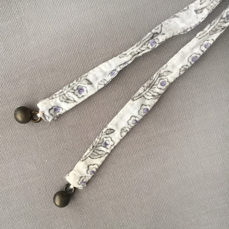May include: Two white fabric cords with a floral pattern and purple accents. Each cord has a silver metal bead at the end.