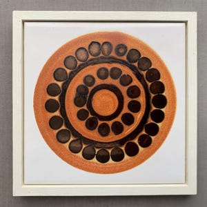 Può includere: Piastrella in ceramica incorniciata con un design circolare. Il design presenta anelli concentrici di punti e linee marroni e marrone scuro su sfondo arancione. La piastrella è inserita in una cornice di legno bianco, creando un'opera d'arte da parete decorativa.