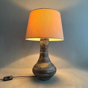 Könnte beinhalten: Tischlampe mit einem braunen, strukturierten Keramikfuß und einem pfirsichfarbenen Lampenschirm. Die Lampe hat ein klassisches, elegantes Design mit Kabel und Schalter. Der Schirm hat eine warme Farbe. Die Lampe ist etwa 40 cm hoch.