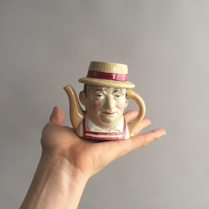May include: A ceramic teapot shaped like a person's head wearing a straw hat with a red band. The teapot has a painted face with a moustache and a red and white striped shirt.