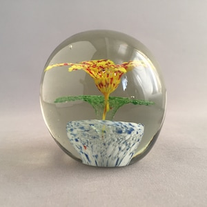 Könnte beinhalten: Ein Glas-Papiergewicht mit einer gelben, roten und grünen Blume im Inneren. Die Blume sitzt auf einem blauen und weißen Sockel.