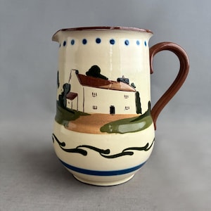以下が含まれることがあります： クリーム色のセラミックピッチャー。茶色のハンドルと縁が付いています。白い家と茶色の屋根、緑の木々、茶色と緑のベースの手描きの風景画が特徴です。青い点と青いストライプが上部と下部を飾っています。
