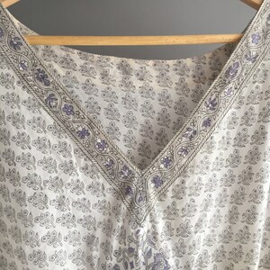 May include: A white cotton top with a V-neckline and a floral print in shades of grey and purple. The neckline is trimmed with a decorative border.