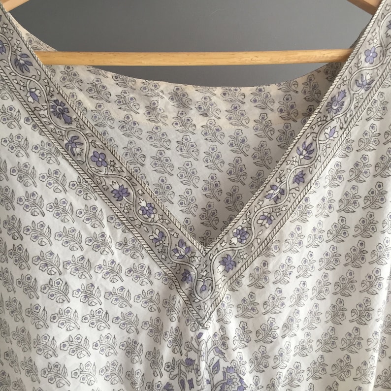 May include: A white cotton top with a V-neckline and a floral print in shades of grey and purple. The neckline is trimmed with a decorative border.