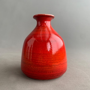Può includere: Un vaso in ceramica rosso vivo con finitura lucida. Il vaso ha un corpo arrotondato e un collo stretto, con un bordo leggermente svasato. La trama del vaso sembra avere striature orizzontali. Lo sfondo è grigio.