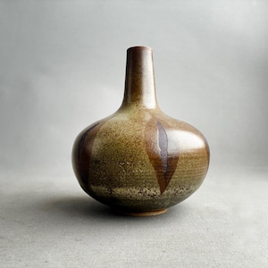 Può includere: Un vaso in ceramica fatto a mano con un corpo bulboso e un collo sottile. Il vaso presenta una glassa screziata verde e marrone con accenti verticali e a forma di freccia marrone scuro. La base è di un colore più chiaro, non smaltato.