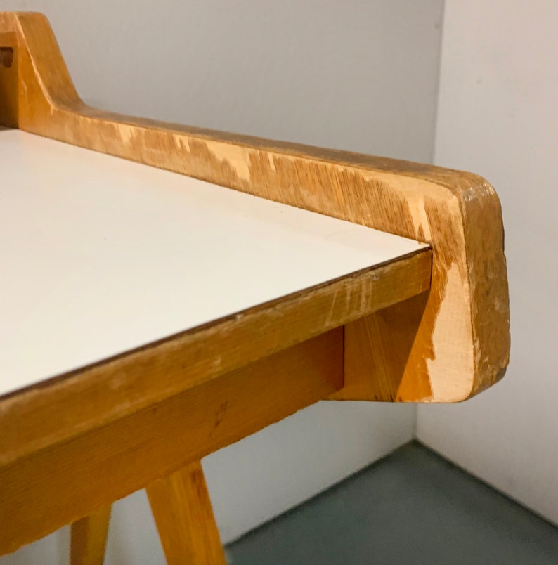 May include: Close-up of the corner of a wooden table with a white laminate top. The table has a simple design with a slightly angled top edge and a visible wood grain.