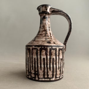 Può includere: Brocca in ceramica con manico, caratterizzata da una smaltatura maculata marrone e bianca. La parte inferiore presenta un motivo geometrico verticale marrone scuro. La brocca ha un collo stretto e un bordo leggermente svasato.