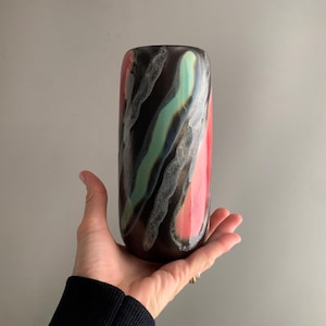 Può includere: Un vaso cilindrico con una base marrone scuro e striature astratte colorate. Le striature includono rosso, verde e bianco, creando un motivo unico. Il vaso è tenuto in una mano, mostrando le sue dimensioni e il suo design.