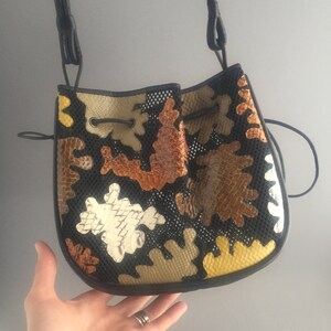 May include: A black bucket bag with a drawstring closure. The bag is made of a black mesh fabric and features a patchwork design of brown, tan, and white leather pieces. The bag has a long black strap.