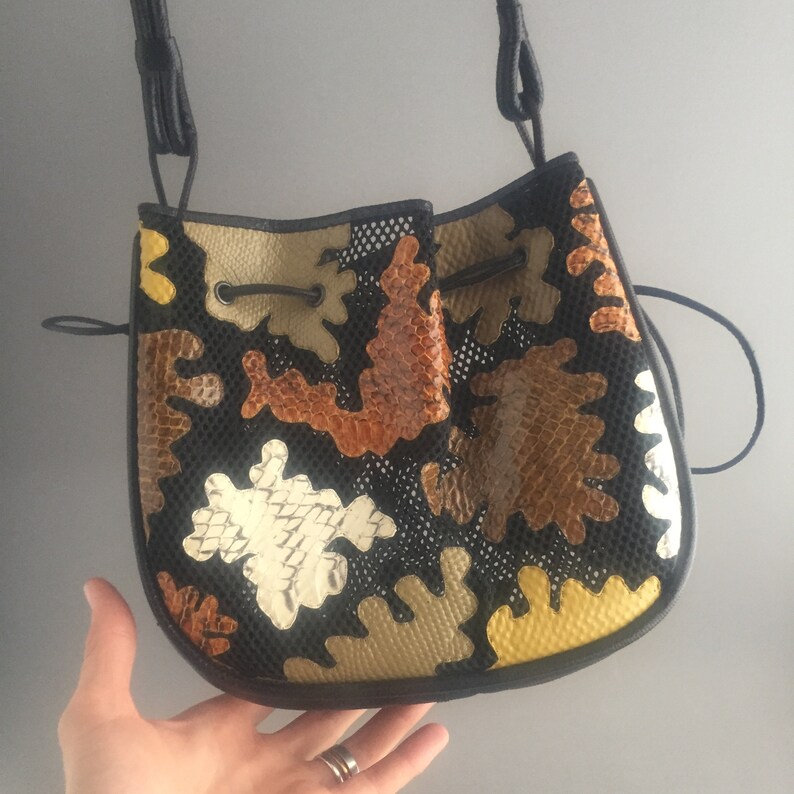 May include: A black bucket bag with a drawstring closure. The bag is made of a black mesh fabric and features a patchwork design of brown, tan, and white leather pieces. The bag has a long black strap.
