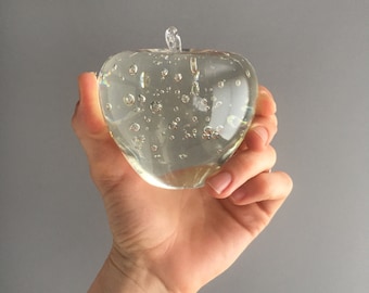 Könnte beinhalten: Ein durchsichtiger Glas-Apfel-Briefbeschwerer mit kleinen Luftblasen. Der Apfel ist herzförmig und hat eine glatte, polierte Oberfläche.