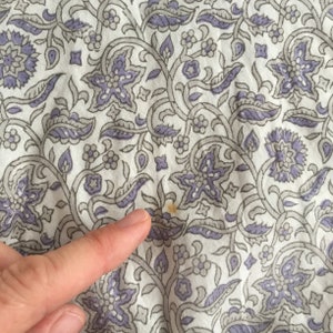 May include: White fabric with a repeating pattern of grey and purple floral designs. There is a small brown stain on the fabric.