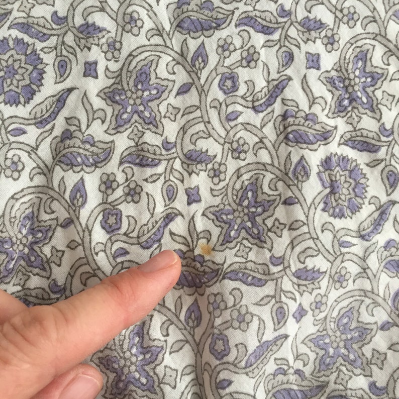 May include: White fabric with a repeating pattern of grey and purple floral designs. There is a small brown stain on the fabric.