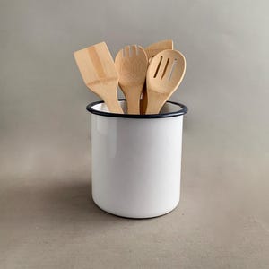 May include: White enamel utensil holder with a black rim, filled with four wooden kitchen utensils, including a spatula, a slotted spoon, and two spoons.