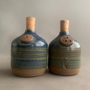 Puede incluir: Un juego de dos saleros y pimenteros de cerámica azul y marrón con tapones de corcho. Los saleros tienen un patrón a rayas y una parte superior con forma de botón con agujeros para dispensar sal y pimienta.