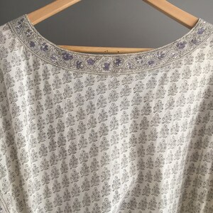 May include: White cotton fabric with a delicate floral print in shades of grey and purple. The fabric has a scalloped neckline with a decorative trim.